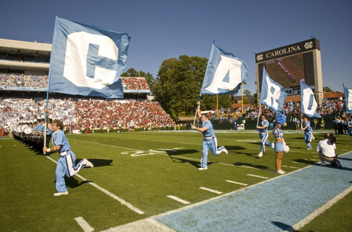 Male cheerleaders running on football field with flags spelling out Carolina, only C, A, R, and O are visible