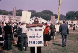 Black man with glasses holds sign that says CIVIL RIGHTS PLUS FULL EMPLOYMENT EQUALS FREEDOM in front of crowd of people holding signs.