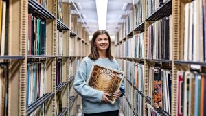 Caroline Norland Has light skin and shoulder-length brown hair. She stands in the Music Library, wearing a gray sweatshirt and holding a recording of Handel: Overtures.