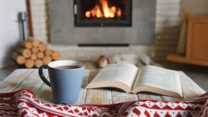 Color photo of a cozy scene in front of a lit fire in a fireplace. An open book sits on a coffee table beside a cup of coffee and a soft blanket.