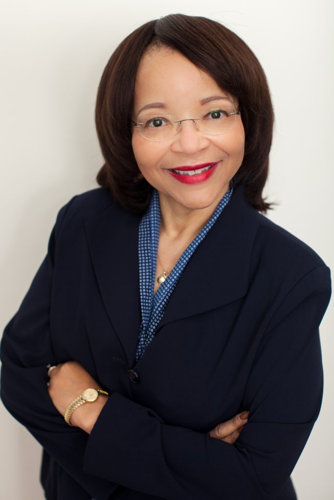 Maxine Brown-Davis smiles for a portrait with her arms crossed. Maxine wears a dark navy sport jacket and a blue blouse. Maxine wears glasses, red lipstick and has shoulder-length straight brown hair.