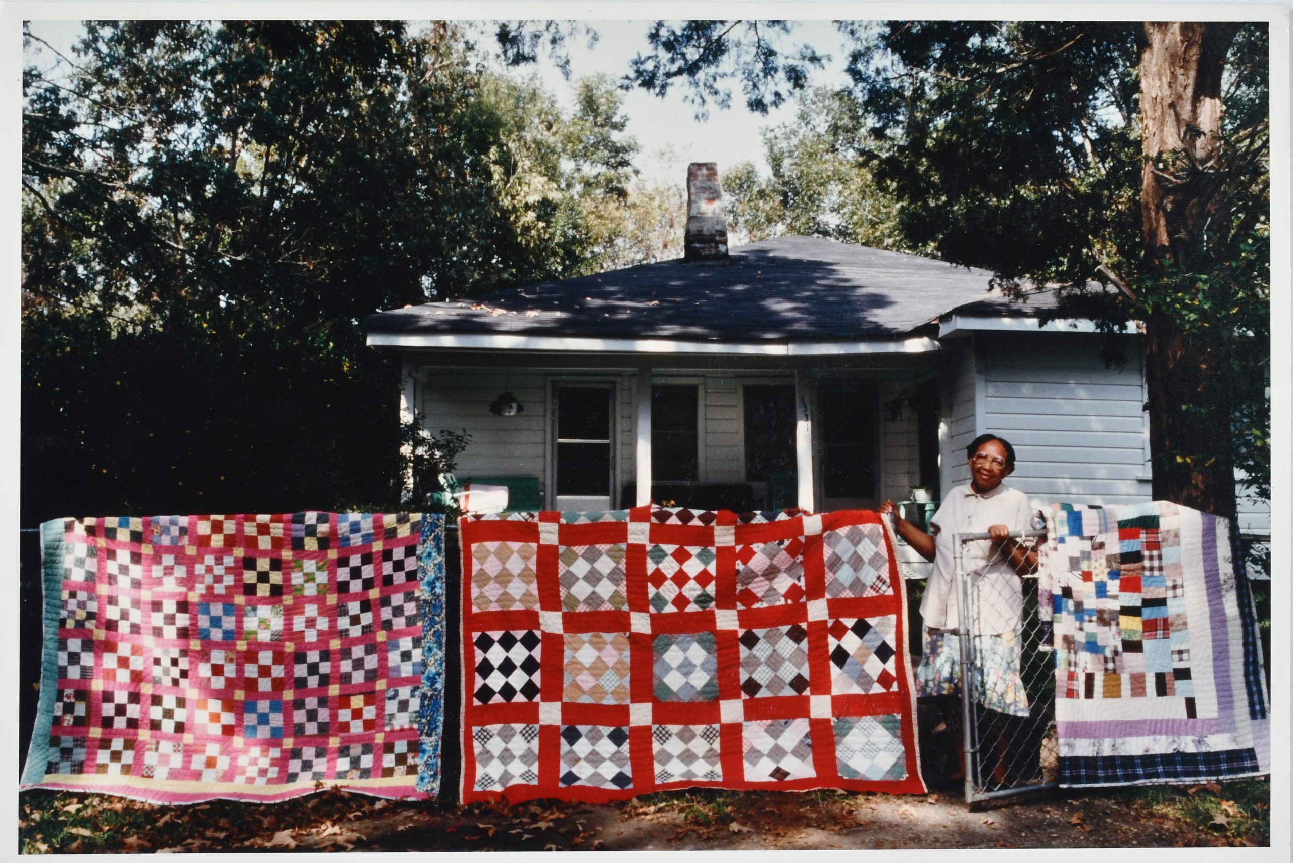 Color archival photograph from the Roland L. Freeman collection in Wilson Library. The photograph shows a black woman standing behind a wire fence in front of a house. There are 3 large quilts hanging on the fence.(191 (2) Roland L. Freeman. Florence Barnes. Wilkinson County, Mississippi, October 1992.)