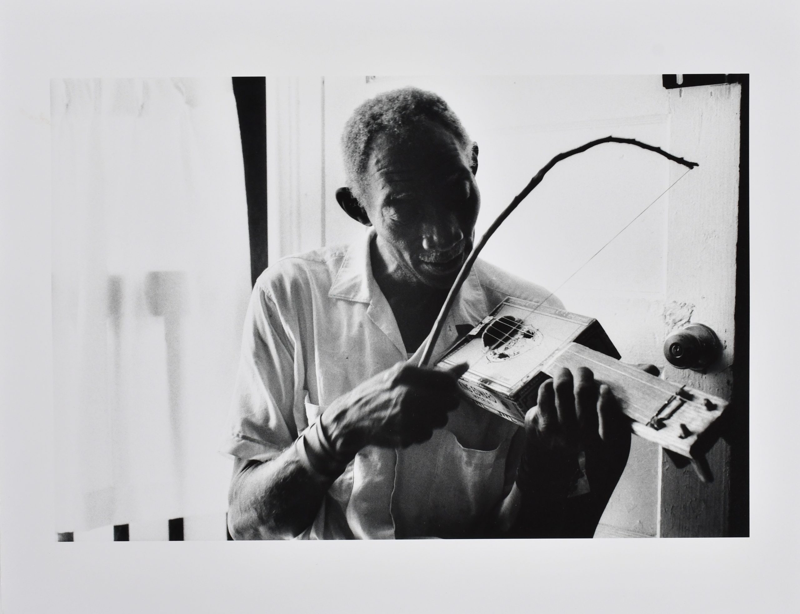Black and white photograph of an older black man playing a cigar box fiddle. (146 (1) Roland L. Freeman, Cigar Box Fiddler Scott Dunbar, Mississippi, 1975.)