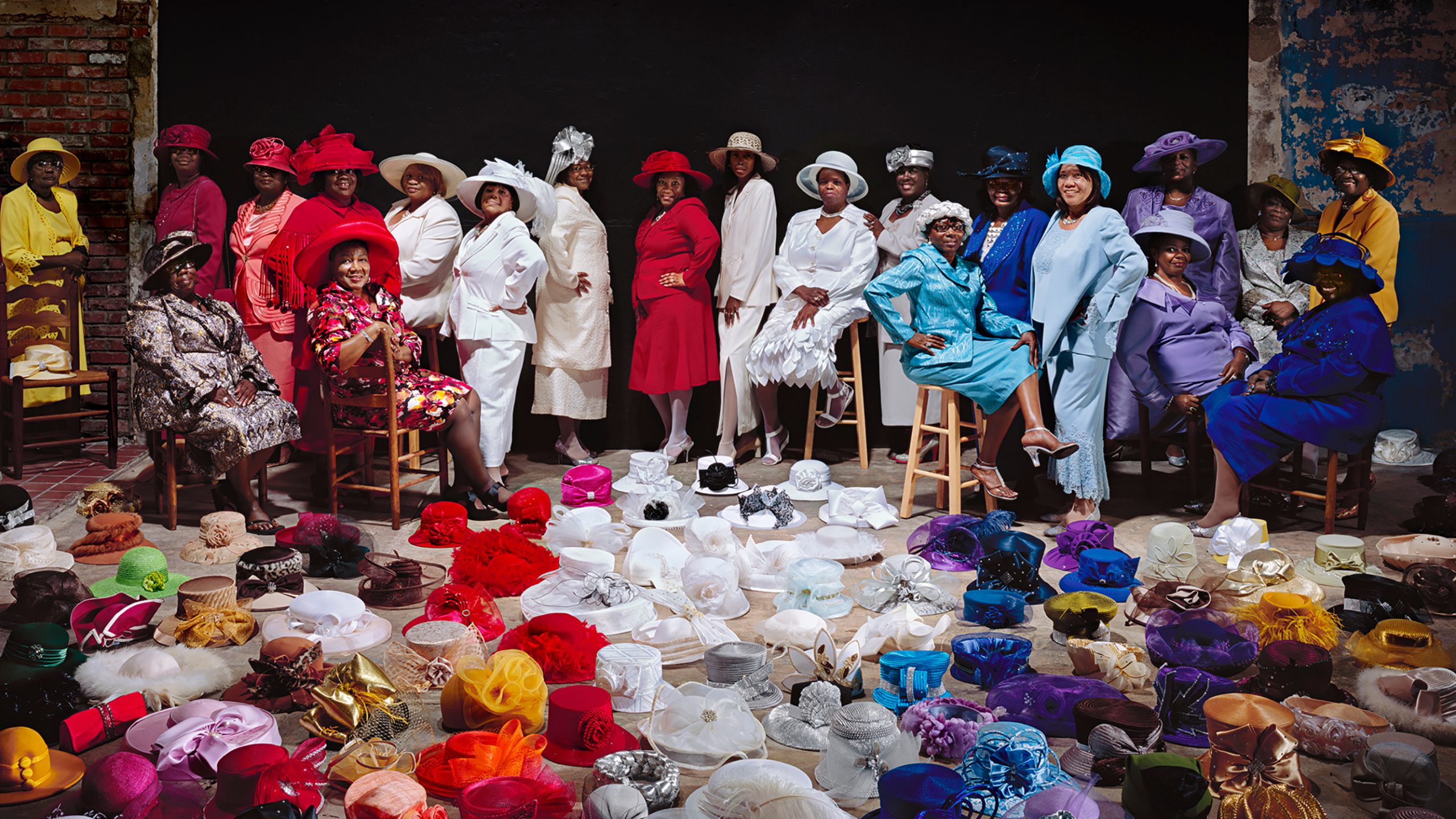 Color photograph of a group of black women dressed in solid colors from the rainbow. In front of the group, is a floor full of colorful hats.