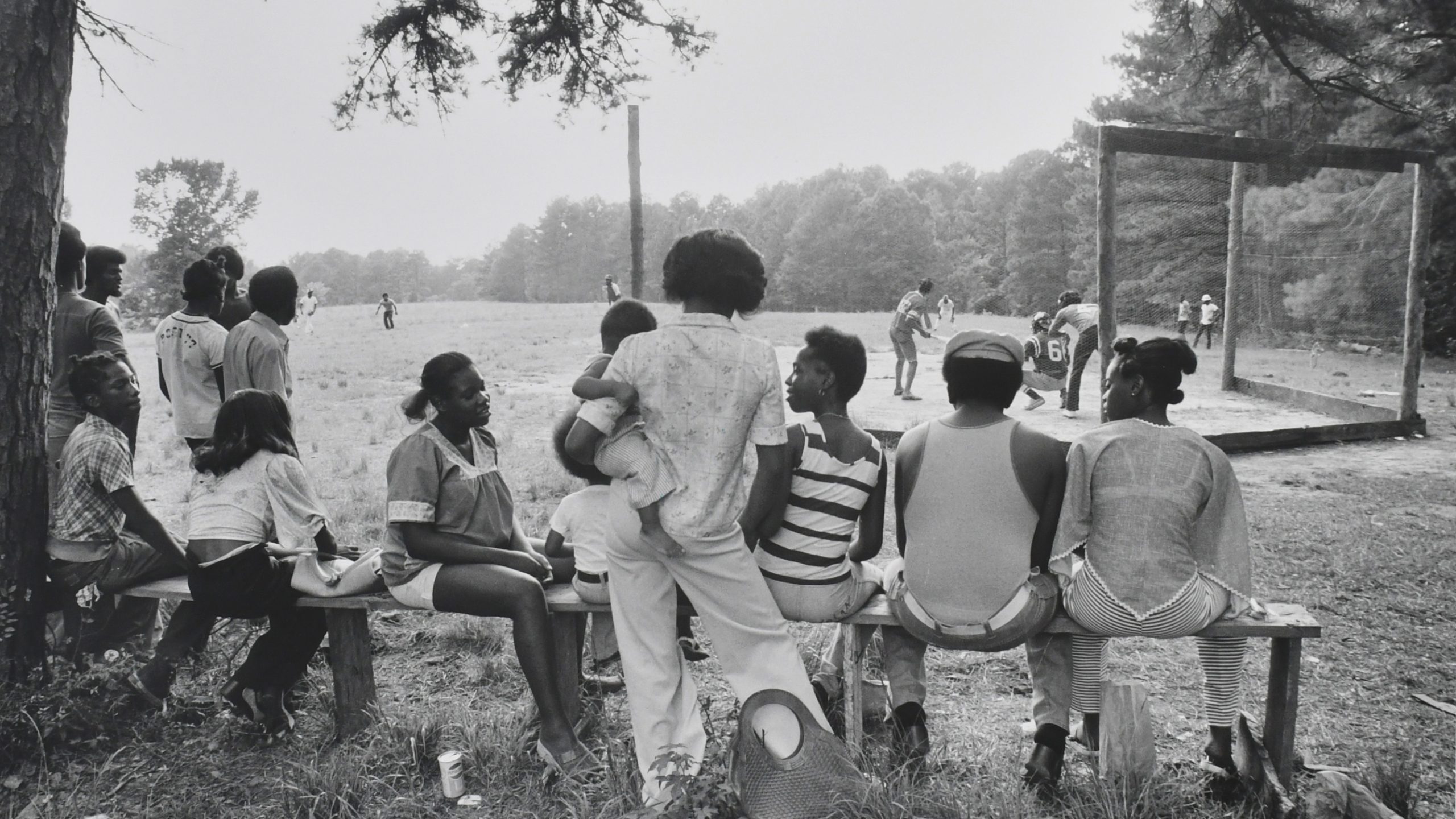 Black and white photograph of a baseball game happening in an open grass field. A crowd of black men and women sitting and gathering around a bench is in the foreground of the photo.