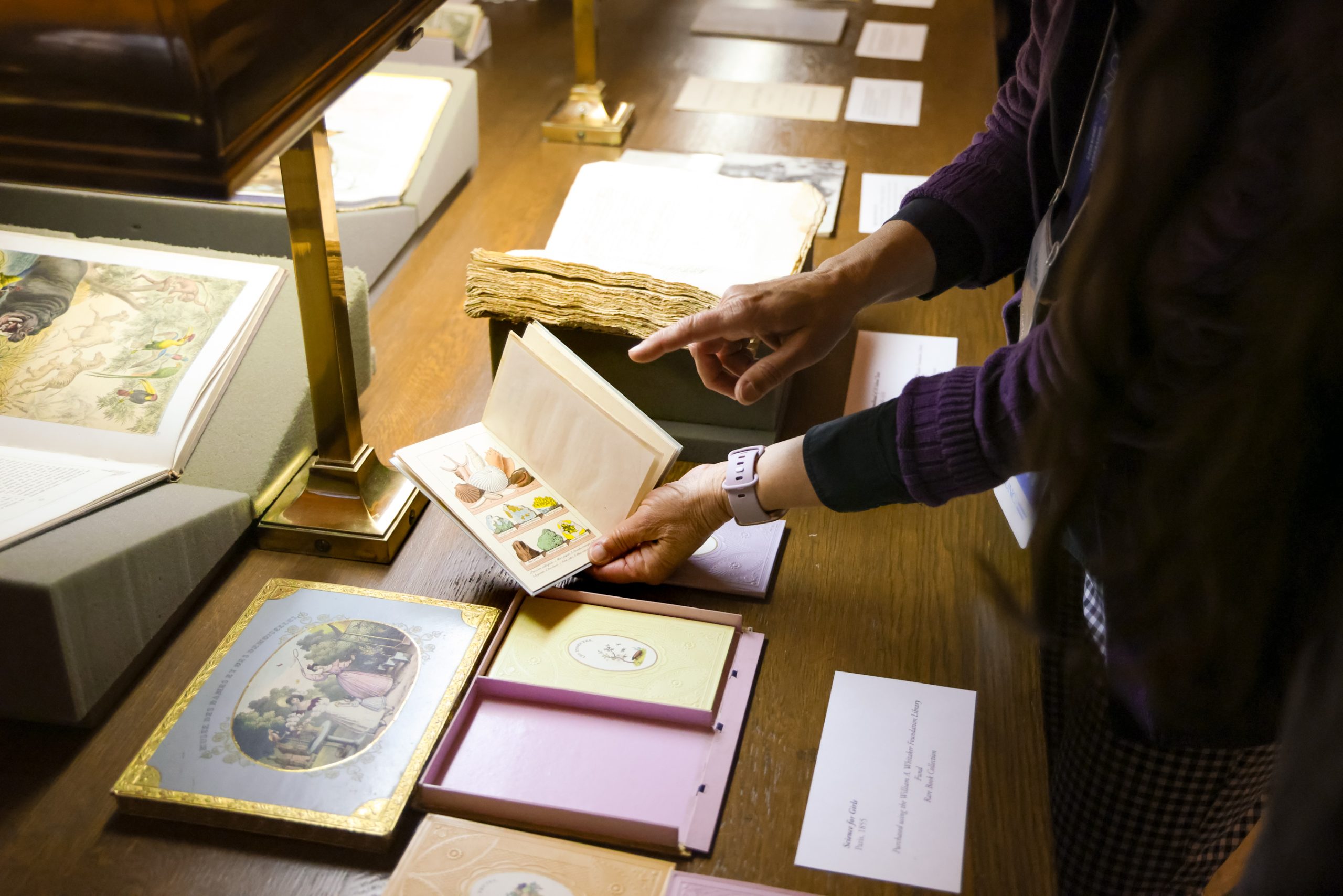 A hand opening a book and pointing. A variety of rare book items are arranged on the table. Recent Acquisitions Evening on April 27, 2023 in Wilson Special Collections Library, UNC-Chapel Hill.