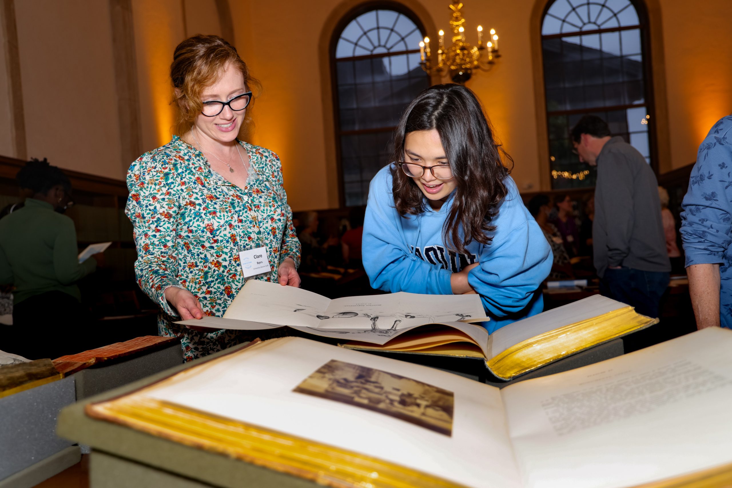 2 young white women lean over large books, examining a paper foldout. Recent Acquisitions Evening on April 27, 2023 in Wilson Special Collections Library, UNC-Chapel Hill.