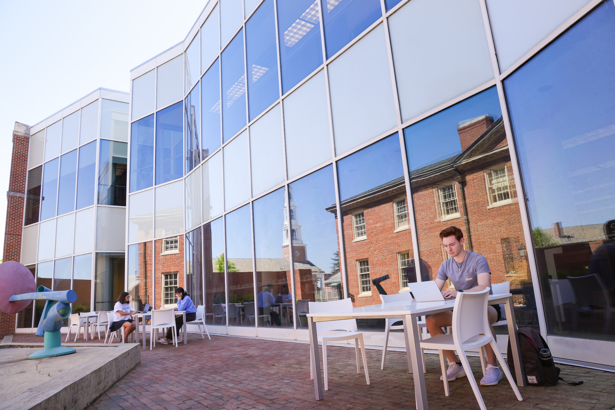 Sloane Art Library, located in Hanes Art Center at UNC-Chapel Hill. Students sitting outside on the Art Library patio space. White tables and chairs are arranged on the brick patio. Huge floor to roof windows of the Hanes Art Center building is in the background; reflecting the blue sky and brick buildings in the distance. 