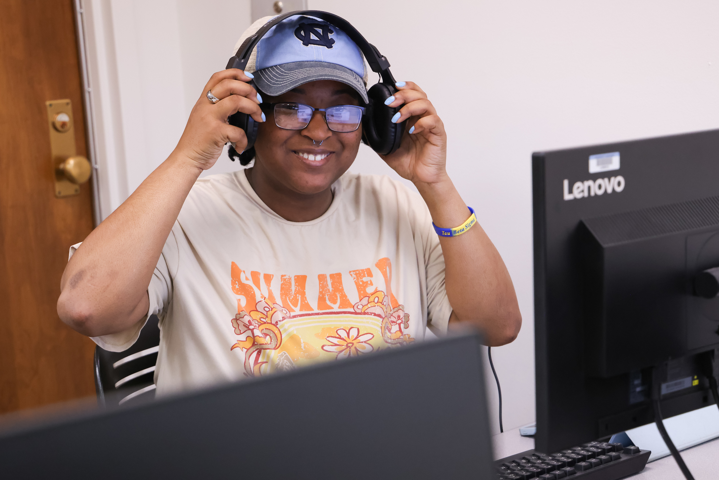 student wearing headphones smiles at the camera, sitting in front of a computer