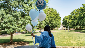 A student sits on a brick wall and looks across a green space on campus. She wears Carolina Blue regalia and holds blue and white balloons, one of which bears the Carolina logo.